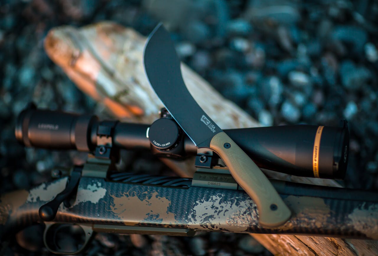 service-01 Detailed shot of a hunting knife resting on a camouflage rifle outdoors.