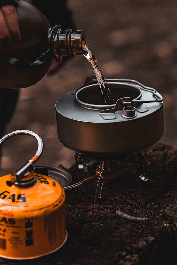 why-choose-us-01 Outdoor camping scene with water being poured into a kettle on a portable gas stove.